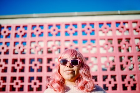A portrait of a cheerful smiling multi-ethnic woman with pink colored hair, standing in front of a pink painted wall.  She puts on pink colored sunglasses.