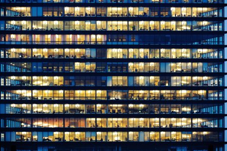 Lots of people working late. Employees seen as silhouettes against their brightly lit offices with large windows. Building framed by the "blue hour" evening sky