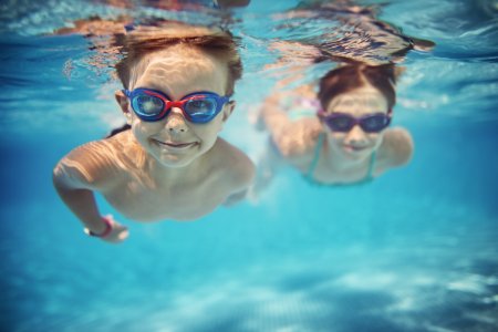 Smiling kids enjoying underwater swim in the pool towards the camera. Sunny summer day. Brother aged 5 is in the front, the sister is aged 9 and is swimming in the background. Slightly soft.