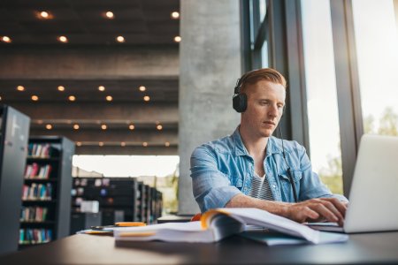 Young male student with headphones studying on the laptop. Handsome caucasian man sitting at the desk and working on laptop at public library.