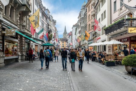 People on shopping street Rennweg with boutique shops, flags on old buildings and St. Peter church clock tower in Zurich, Switzerland.