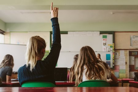 Rear view of female student sitting in the class and raising hand up to ask question during lecture. High school student raises hand and asks lecturer a question.