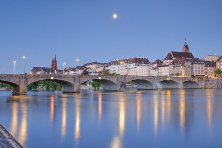 View from the banks of the Rhine along the river promenade to the old town of Basel illuminated at night with the Basel Cathedral, St. Martin's Church, the Mittlere Brücke and the Rhine River, by moonlight