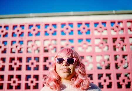 A portrait of a cheerful smiling multi-ethnic woman with pink colored hair, standing in front of a pink painted wall.  She puts on pink colored sunglasses.