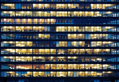 Lots of people working late. Employees seen as silhouettes against their brightly lit offices with large windows. Building framed by the "blue hour" evening sky