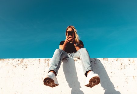 Young woman checking News And Posts On Mobile Phone sitting on modern concrete wall. Simplicity Modern Urban Style. Shot from below against the sky. Urban Lifestyle Youth Portrait.