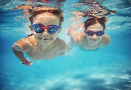 Smiling kids enjoying underwater swim in the pool towards the camera. Sunny summer day. Brother aged 5 is in the front, the sister is aged 9 and is swimming in the background. Slightly soft.