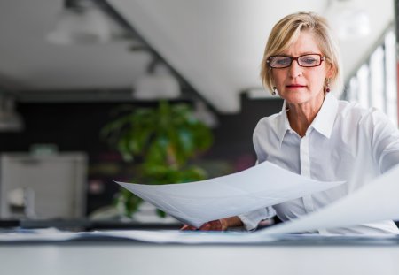 A photo of mature businesswoman examining documents at desk. Concentrated professional is analysing papers in office. Executive is in formals.