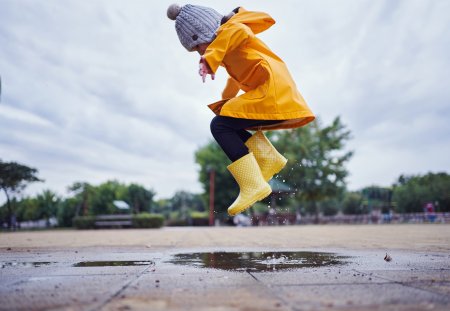 Cute and playful female child jumping in a puddle of water on the street wearing yellow rubber boots and a raincoat.