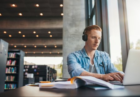 Young male student with headphones studying on the laptop. Handsome caucasian man sitting at the desk and working on laptop at public library.