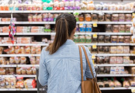 In this rear view, an unrecognizable woman stands with a shopping cart in front of a shelf full of food in the bread aisle of a grocery store.