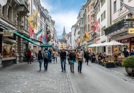 People on shopping street Rennweg with boutique shops, flags on old buildings and St. Peter church clock tower in Zurich, Switzerland.