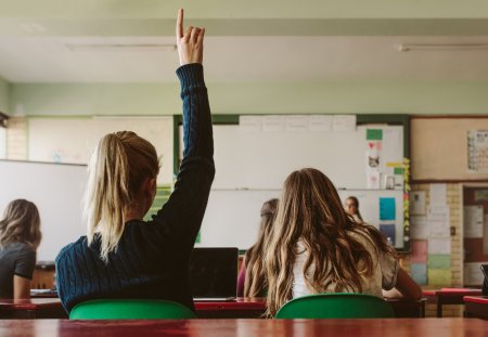 Rear view of female student sitting in the class and raising hand up to ask question during lecture. High school student raises hand and asks lecturer a question.