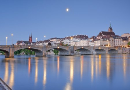 View from the banks of the Rhine along the river promenade to the old town of Basel illuminated at night with the Basel Cathedral, St. Martin's Church, the Mittlere Brücke and the Rhine River, by moonlight