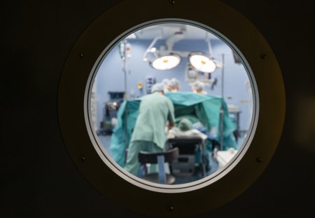 Blurred view into an operating theatre through a porthole in a door in a hospital with unrecognisable people.